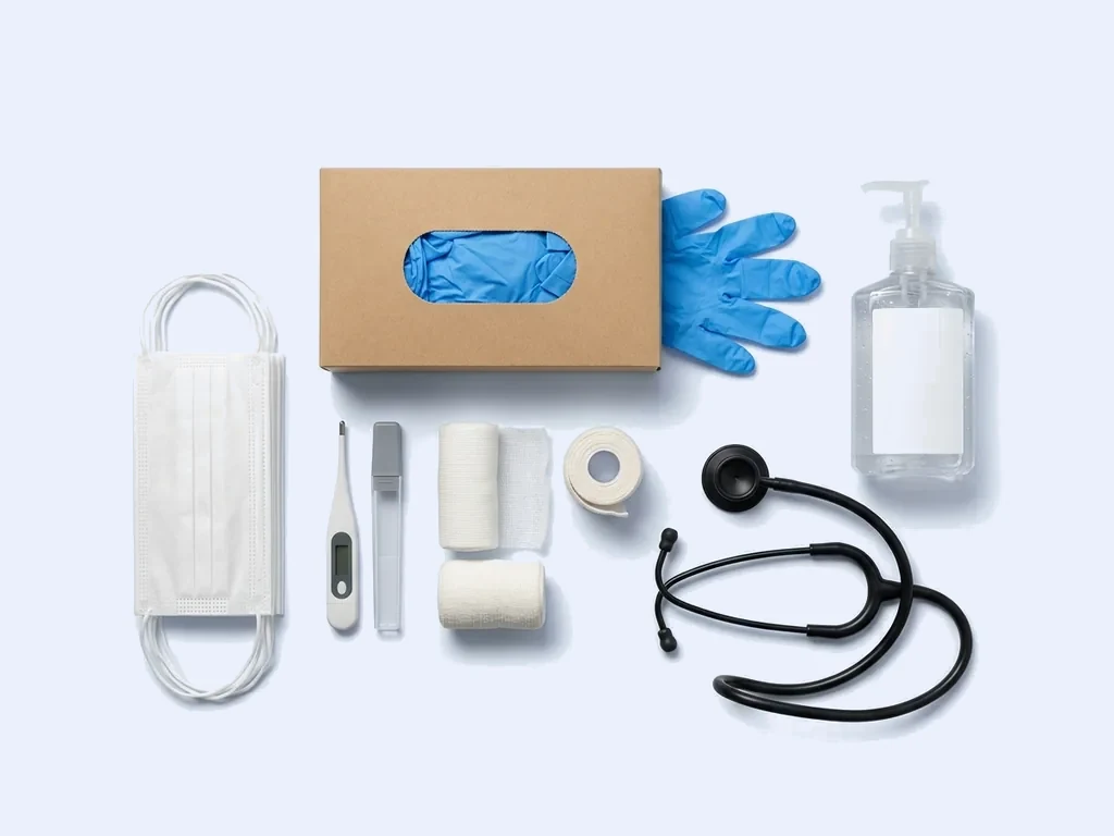 Surgical masks, a digital thermometer, gauze rolls, a box of blue nitrile gloves, a stethoscope and a pump bottle of sanitiser on a pale studio ground